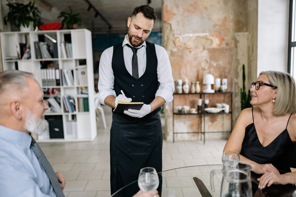 Waiter Taking Order from Customers at a Glass Table in an Elegant Interior with Textured Walls